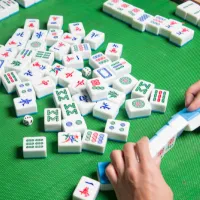 Mah Jongg tiles spread across a green playing surface, with a player's hands organizing tiles on a rack.