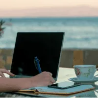 A person writing in their notebook next to their laptop on a beachfront.