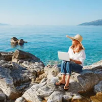 A woman sitting on a rock along the coastline holding an open laptop.