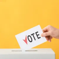 image of a hand holding a ballot that says vote placing it into a box with yellow background