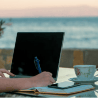 A person writing in their notebook next to their laptop on a beachfront.