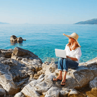 A woman sitting on a rock along the coastline holding an open laptop.
